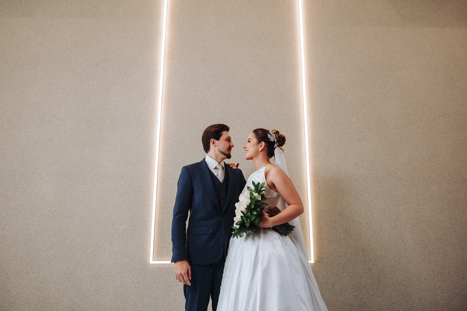 Noivos com buquê diante de uma parede clara com linhas de luz